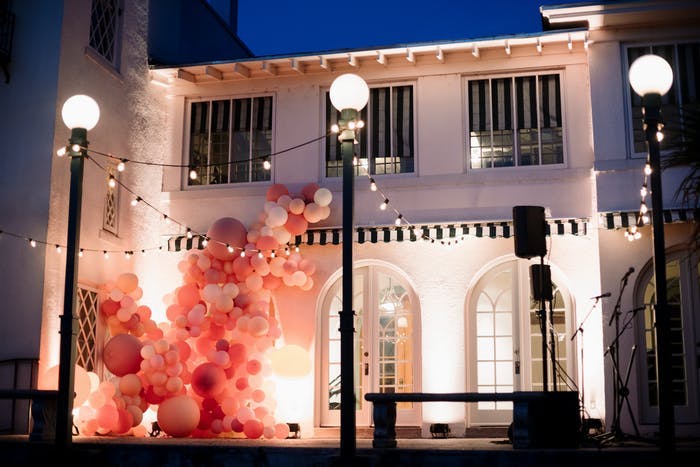 A white building with three lamp posts in front. Pink and coral balloon installations look as if they are growing on the building like ivy