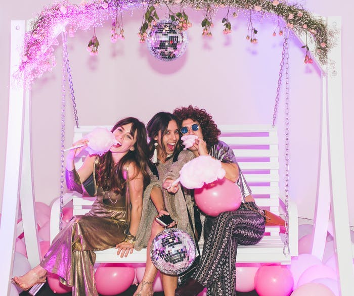 Three girls sit in a swinging bench with a pink backdrop holding disco balls and eating cotton candy | PARTYSLATE
