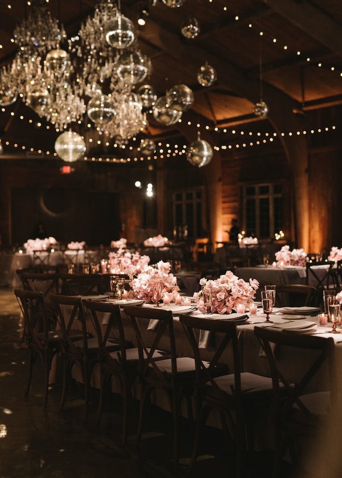 Tables of varying heights with pink florals. In the corner there is a cluster of different sizes disco balls with golden light shining on them | PartySlate
