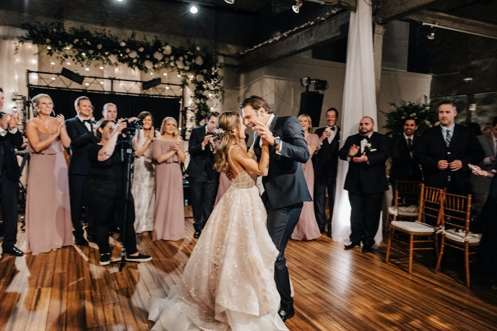 Bride and groom dance, surrounded by loved ones,  in raw industrial space with a backdrop of white roses and greenery.