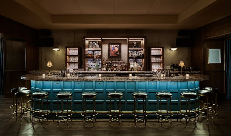 Dimly lit speakeasy bar with wrap around bar with blue side-paneling and leather stools.