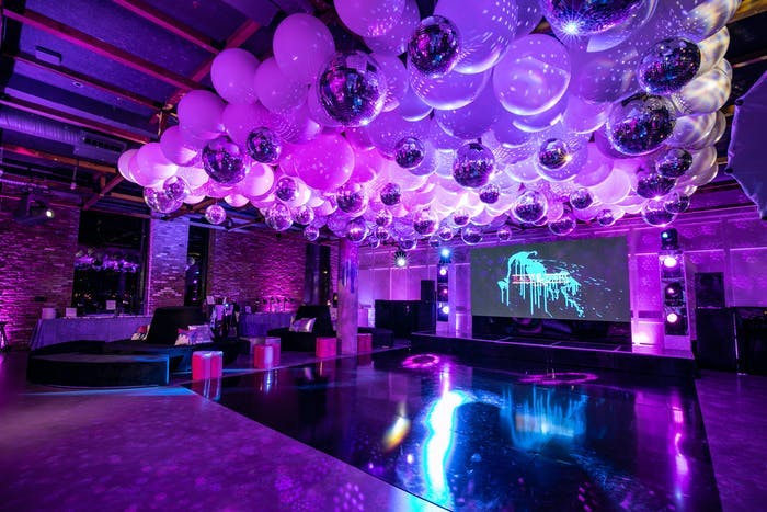 A purple washed room with balloons on the ceiling over the dance floor and a screen displaying blue lighting for disco-themed party | PartySlate
