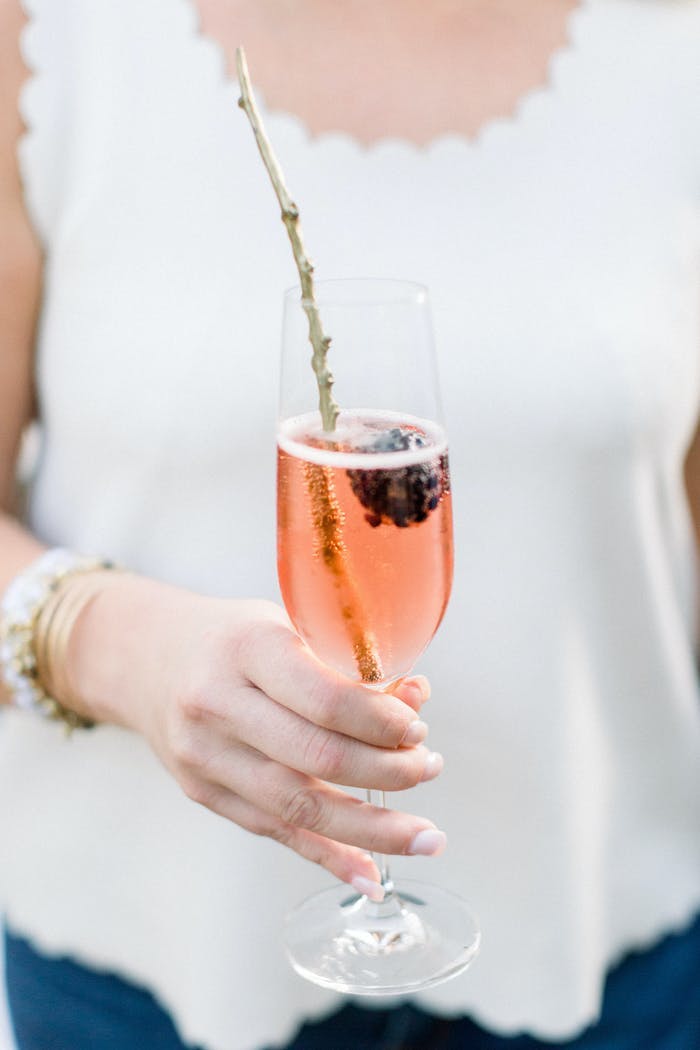 woman in a white tank holding a pink drink in a champagne flute. A branch is used as garnish
