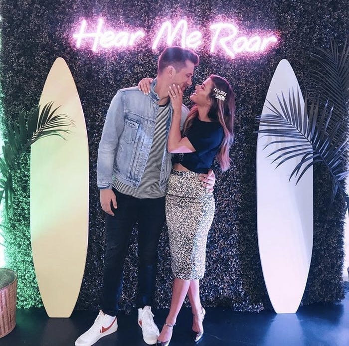 Jojo Fletcher and a man stand face to face between two surfboards and a pink neon sign above them