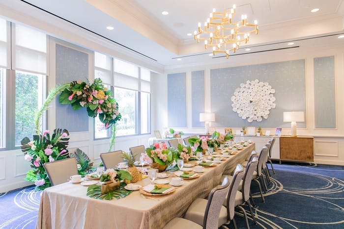 A room with white and powder blue walls. A long table with a beige and white linen and chairs on either side. Clusters of greenery are in the centerpiece and around the room