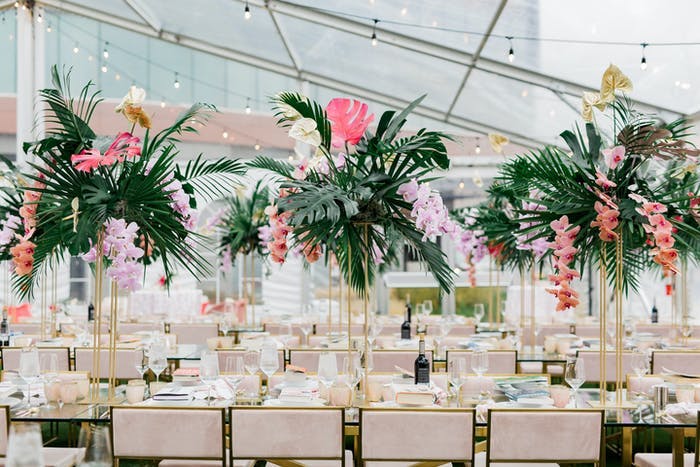 A transparent tend with long tables underneath. Beige chairs along the tables and tall greenery with pink accents as centerpieces.