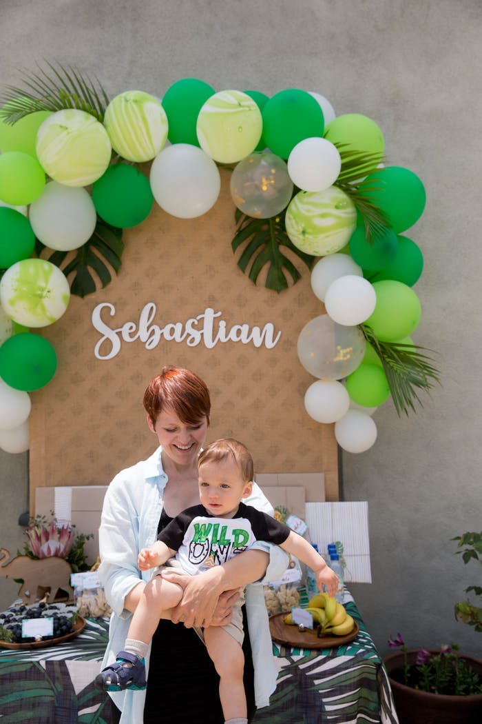 A woman holding a baby in front of a green and brown balloon backdrop outside