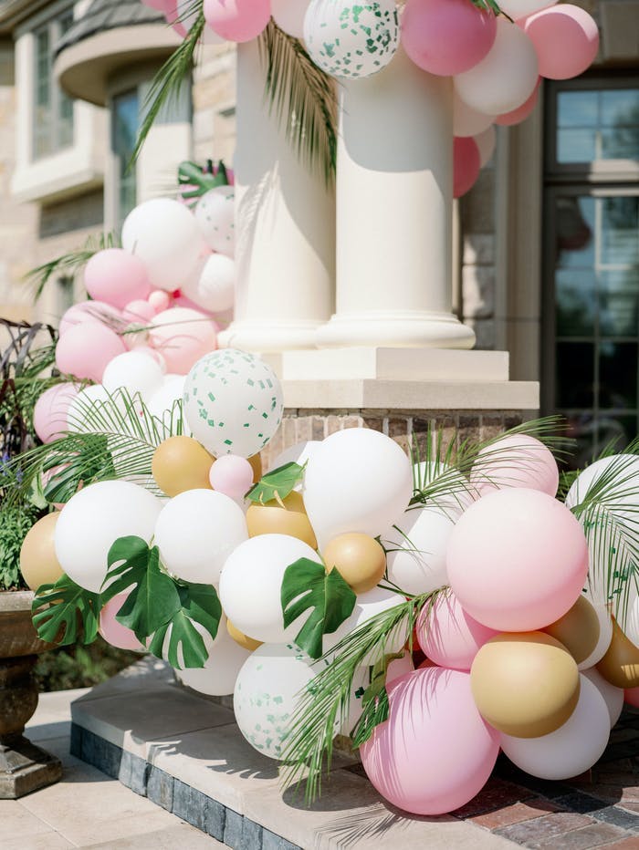 Two columns near the front door of the house surrounded by muted colored balloons in pink, gold, white and silver, and monstera leaves are mixed in