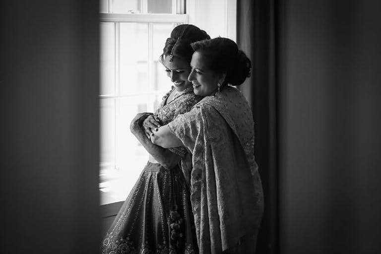 A black and white photo of a mother hugging her daughter from behind in front of a window
