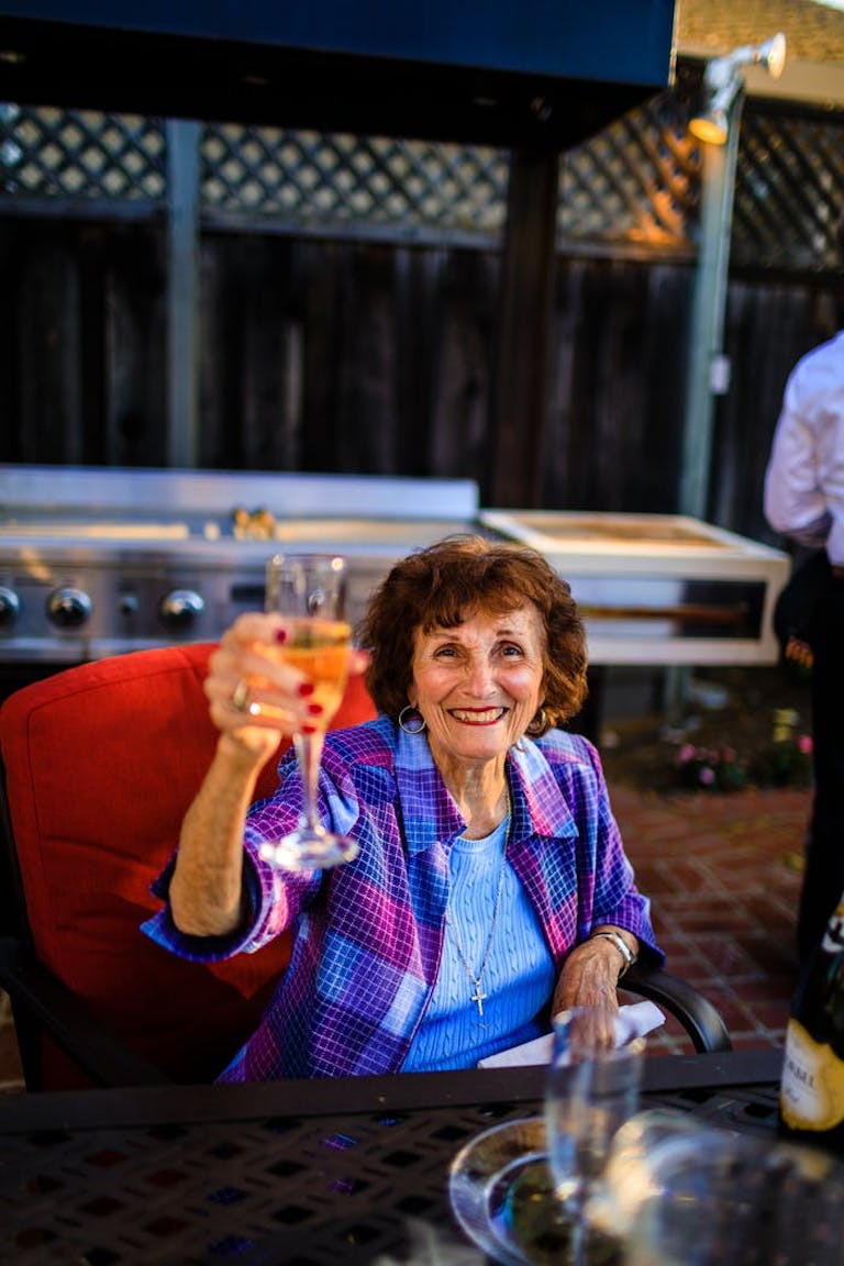 An older woman wearing a blue shirt and purple flannel sits on a red couch and raises a glass of Champagne.