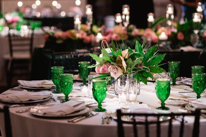 A round table with vintage place settings. Green glass water cups and greenery centerpiece