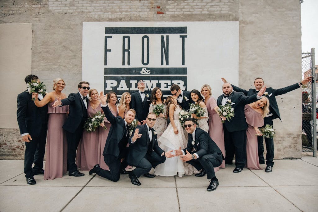 A bride and groom kiss while surrounded by their groomsmen and bridesmaids. The bridesmaids wear rose-colored dresses and hold green and white bouquets. They are all posing in front a light brick wall with a giant black and white sign that reads "Front & Palmer."