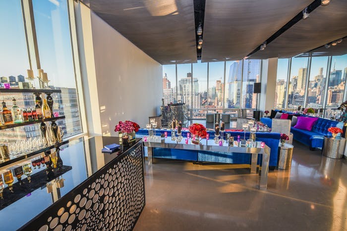 A large room with windows on all sides looking over skylines. Blue couches are in the room with pink pillows and accents