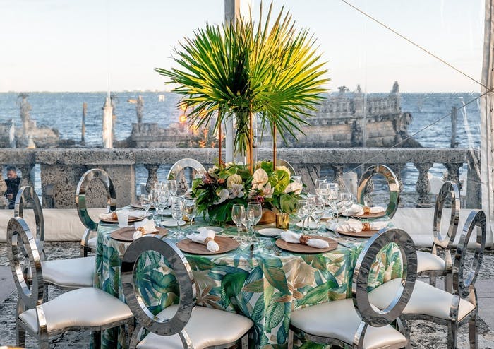 A square table on a rooftop overlooking ruins in the water. Metal chairs surround the table and a green palm leaf is the centerpiece