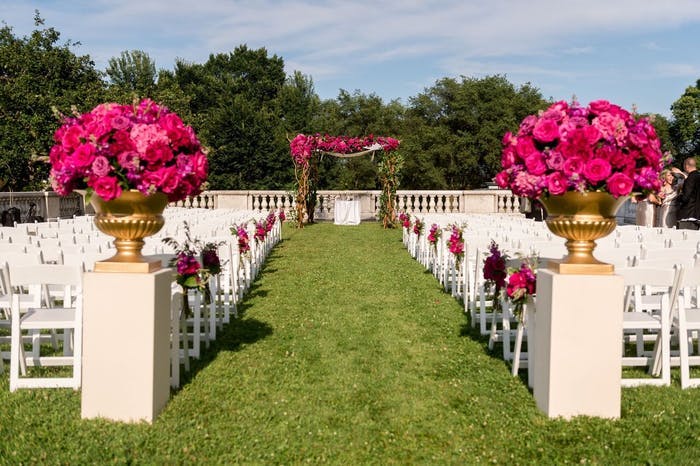 A grassy aisle with white chairs on either side. Pink roses flank the entrance of the aisle.