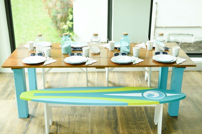 A picnic style table with a wooden finish and place settings. The benches are painted as surf boards