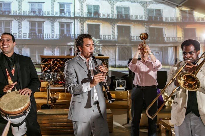 A four piece band on a stage. Two are holding horn instruments and one tilts his head back while playing. Another plays the clarinet wearing a gray suit and the other plays handheld drums