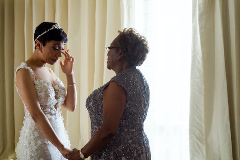 A bride in a white wedding dress wipes away as a tear as her mother holds her hand.