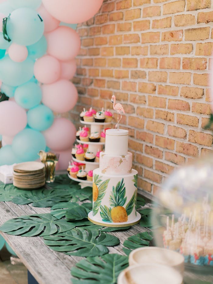 A three tiered cake with pineapples printed on. The table has monstera leaves sitting flat and pink and blue balloons flank the cake