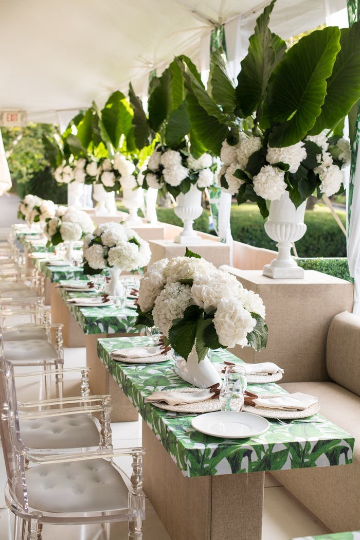 Small individual tables with green and white linens. Translucent chairs surround the tables and tall green and white potted plants are between each table