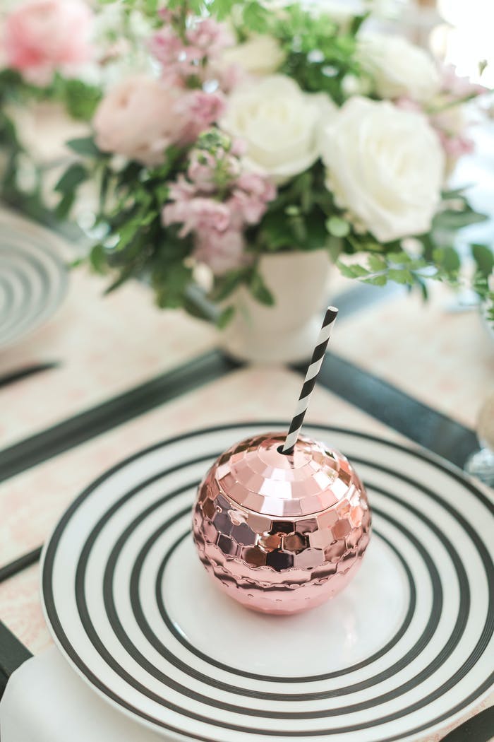 A pink disco ball cup wth a straw sits on a black and white plate. Pink and white florals are in the corner. | PartySlate