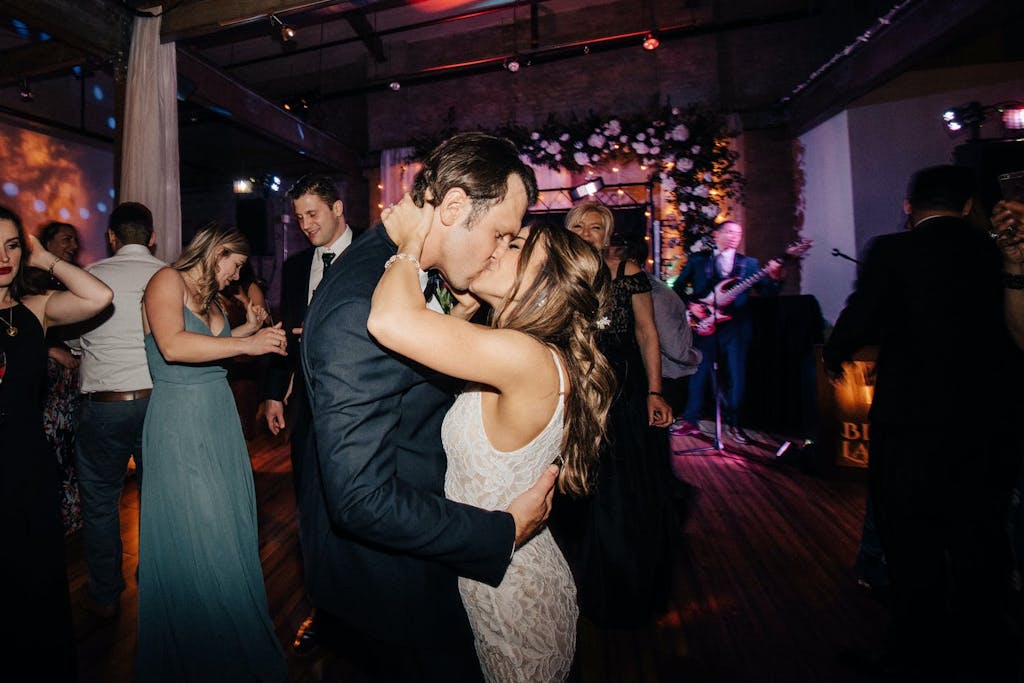 A closeup of a bride and groom kissing with their friends and band performing in the backdrop.