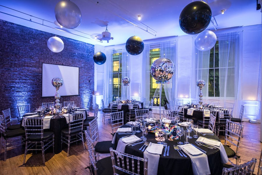 An industrial style room lit up blue. Centerpieces are raised disco balls and balloons are on the ceiling