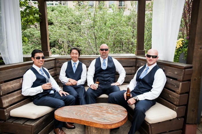 four men wearing white shirts, vests and sunglasses sitting in a semi circle wodden bench.