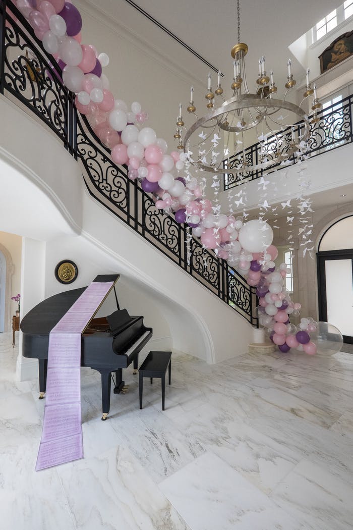A grand staircase with a black piano underneath. A purple runner goes over the piano while pink and white balloons follow the rail up the stairs