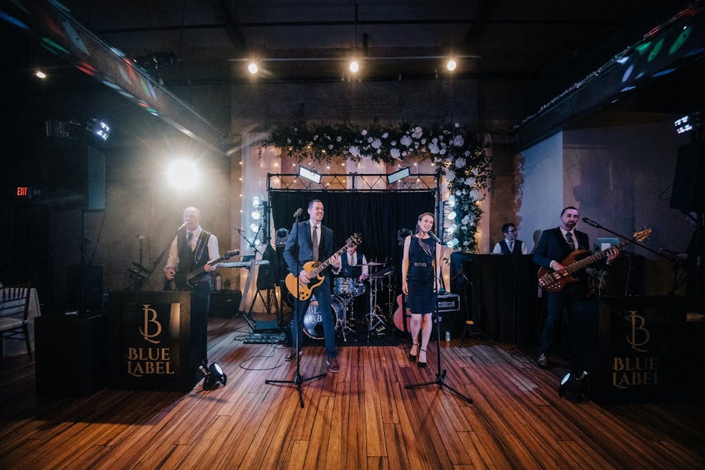 A six-piece band performs on a wooden stage in a raw industrial setting with a backdrop of greenery and white roses.