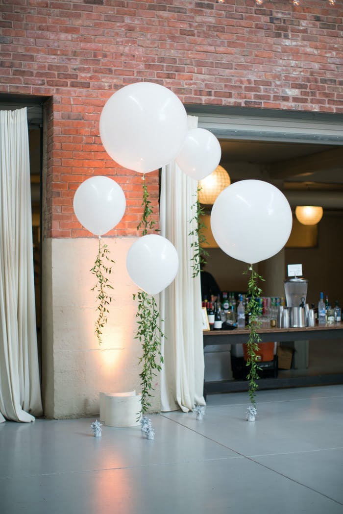 White circular balloons with greenery ribbons against a brick wall and a doorway to a larger table