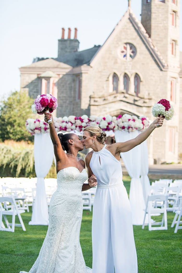 Two brides kiss and hold up their bouquets in front of their aisle and venue