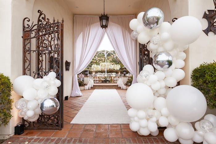 An entrance way with a decorative rod iron gate and terra-cotta tiled floors. White drapes at the end of the entrance and clusters of white balloons at the front