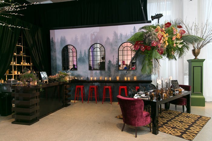 A room with a backdrop of windows and a night sky. A red chair sits at a narrow table with a tall centerpiece thats colorful and green. A bench sits along the backdrop with glowing candles