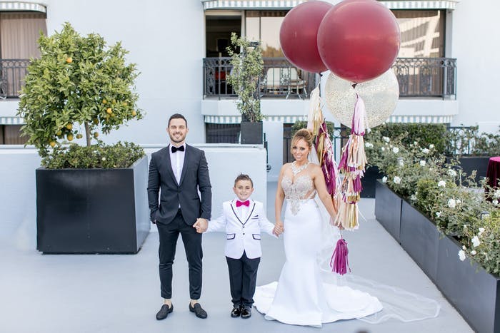 A bride and groom holding hands with their son, the bride holds two maroon balloons with pink tassels as the ribbon
