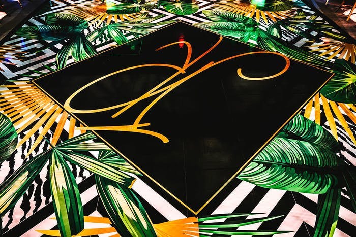 A black square dance floor with gold calligraphy through it. The outside of the dance floor is filled with black and white stripes and banana leaves