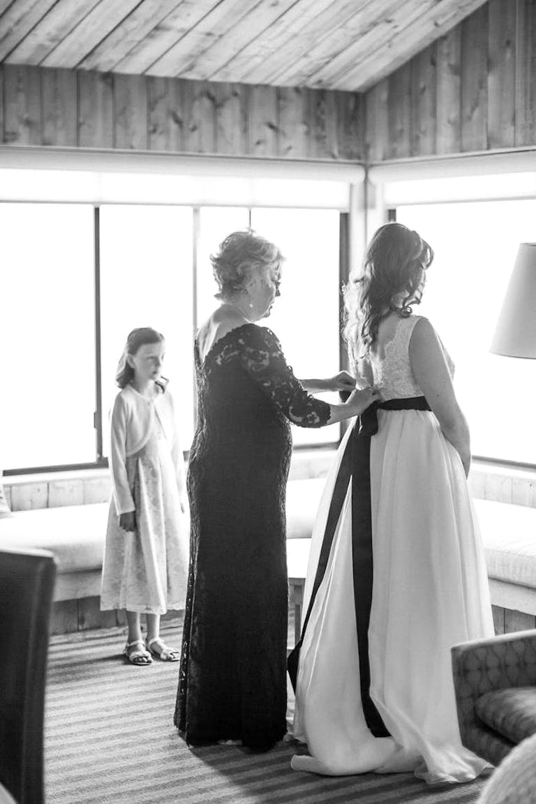 A mom ties the black bow of her daughter's wedding dress while a young girl in a white dress looks on.