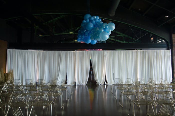 an industrial style warehouse with low white curtains separating the space, transparent chairs sit behind it and a cluster of blue balloons is above the curtain