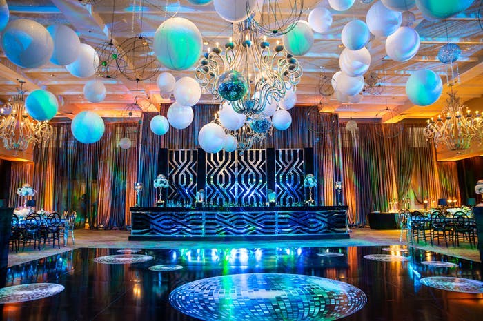 A black dance floor with disco balls printed on it. Multicolored balloons are over the dance floor with a stage in front | PartySlate