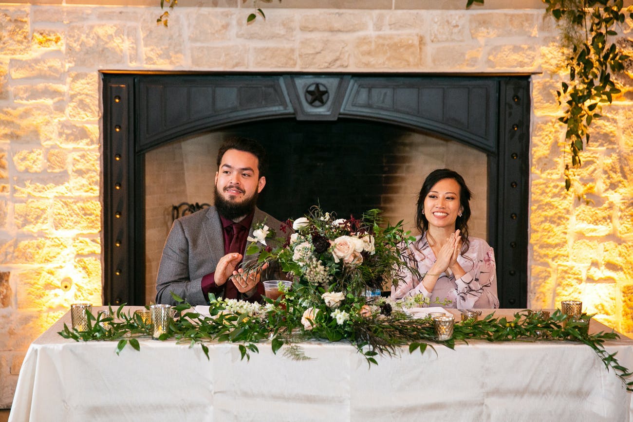 Couple sit at a sweetheart table in front of an uplit fireplace.