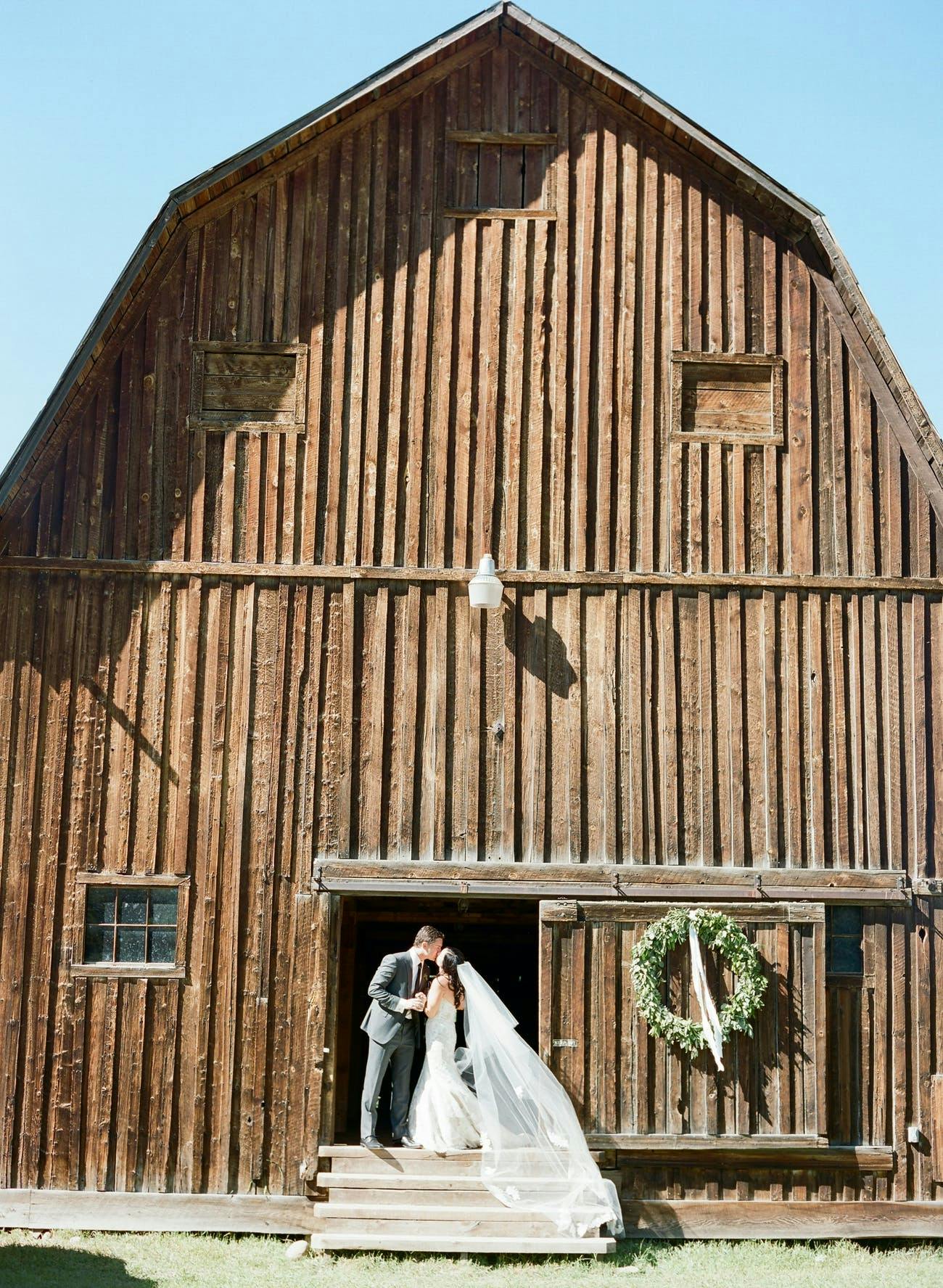 Bride and groom kiss near rustic barn for wedding photo op | PartySlate
