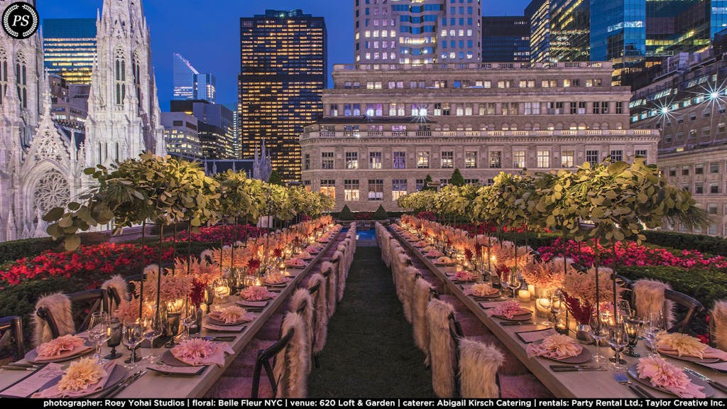 two long tables with raised greenery as the centerpiece and glowing candles below. Pink fur chair throws on each setting and a lit skyline in the background. 