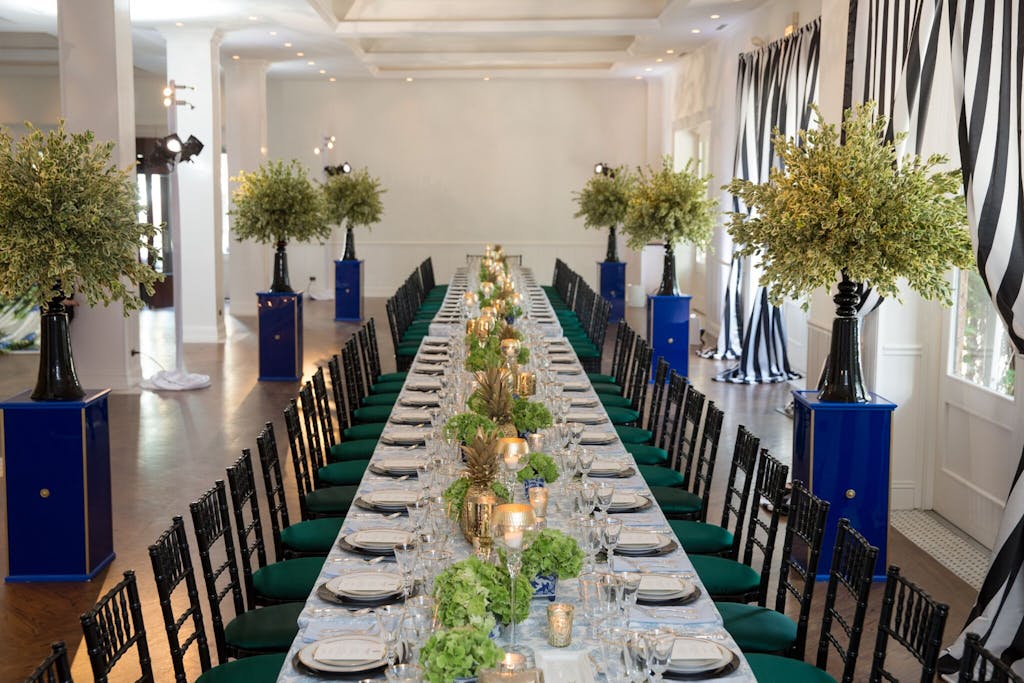 a long table with emerald green chairs on either side and candles and greenery down the middle. White plates and translucent glasses at each place setting and trees on blue pedestals flanking the table. 