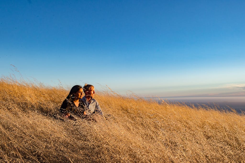 Engaged Couple Sit in Golden Field Under Deep Blue Sky | PartySlate
