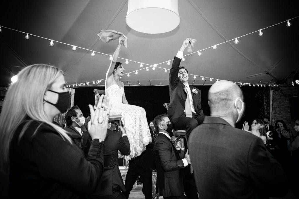 A Woman and Man Wear Masks as The Bride and Groom Are Raised on Chairs During the Horah | PartySlate