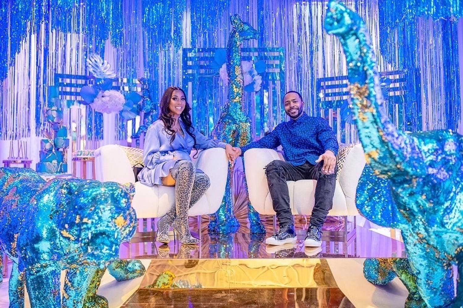 Parents-to-Be Sit on White Chairs Against Iridescent Backdrop and Glittery Blue Animal Sculptures | PartySlate