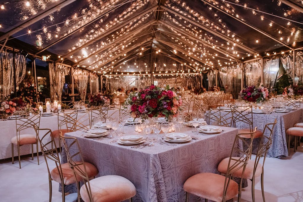 Clear Wedding Tent With String Fairy Lights, Pink Seating, and Pink Flower Centerpieces | PartySlate