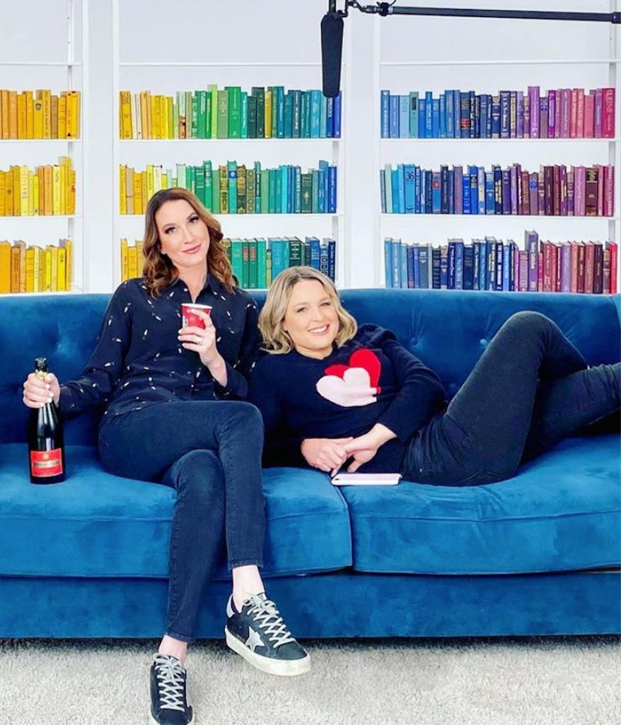 Clea and Joanna from the Home Edit on a blue couch in front of a rainbow organized book shelf