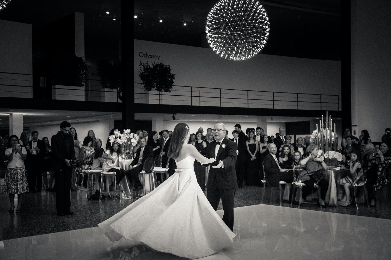 Black and White Photo of Bride Dancing With Father Beneath a Globed Geometric Chandelier | PartySlate