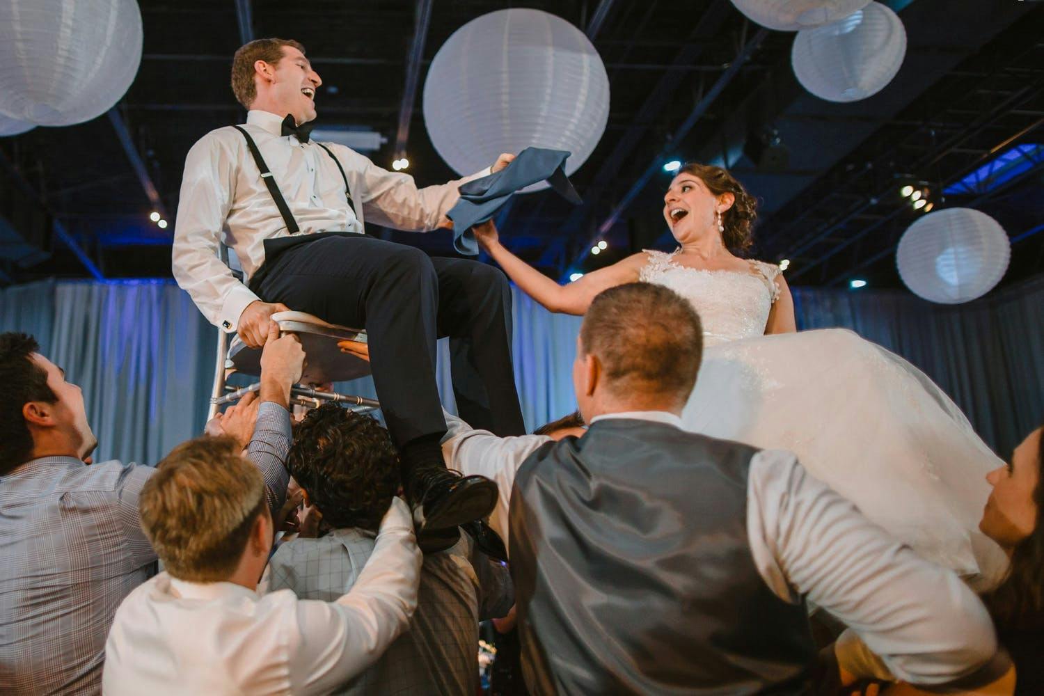 Wedding Hora With Bride and Groom Lifted into the Air Underneath White Globed Paper Lanterns | Partyslate